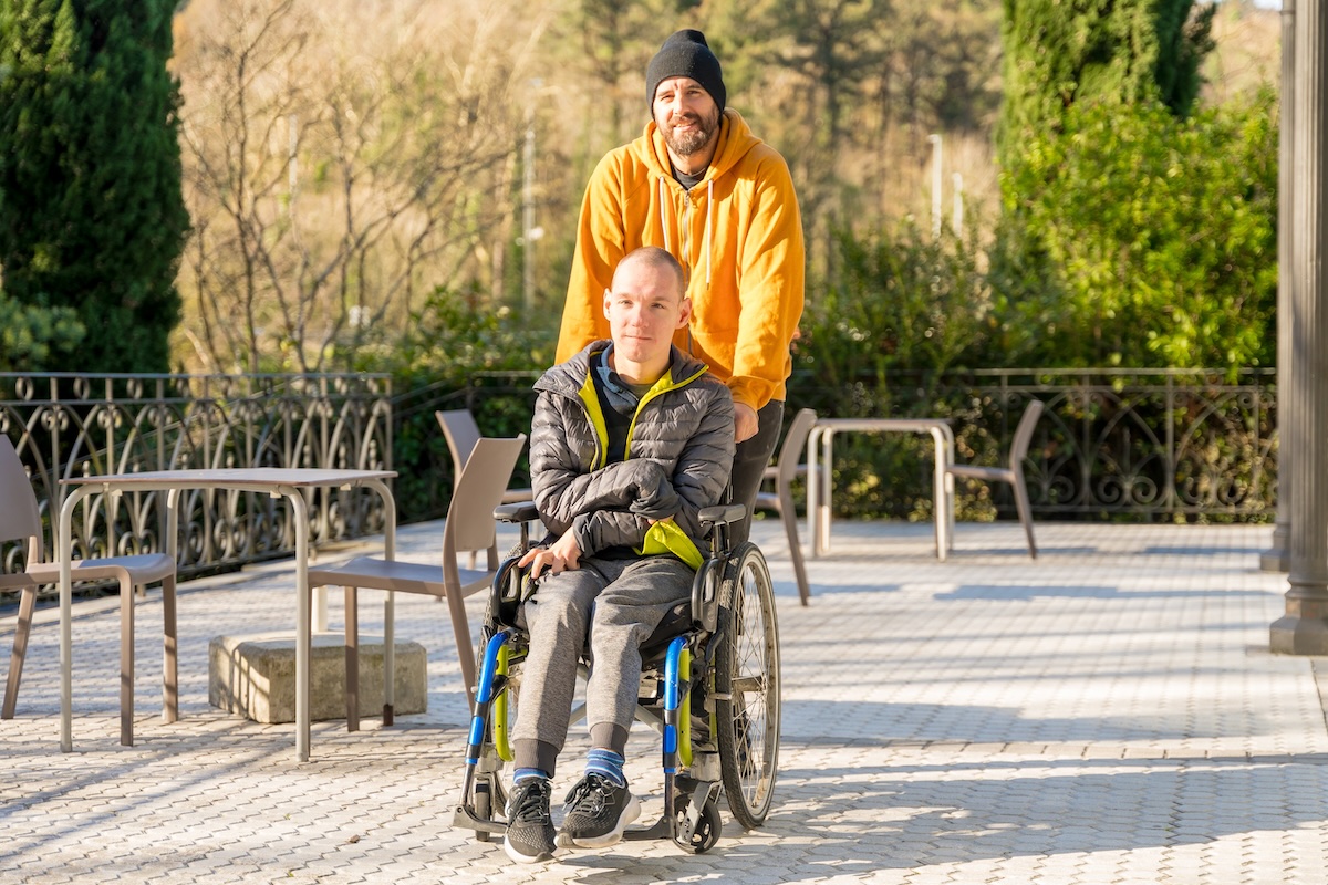 Man on an outdoor deck pushes along his friend who uses a wheelchair.